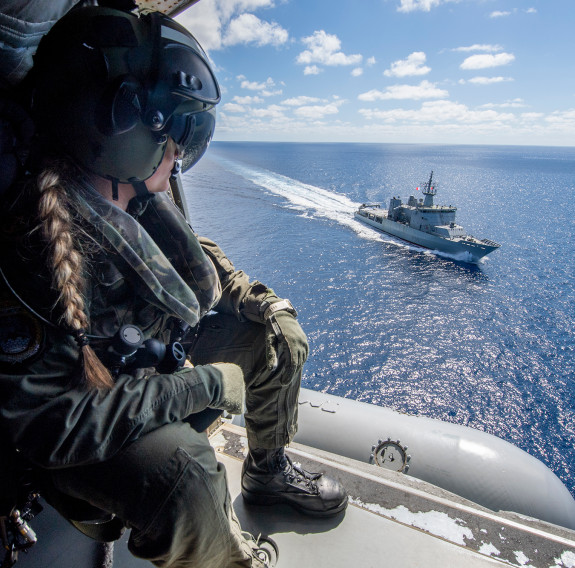 A Royal New Zealand Navy helicopter loadmaster looks out the SH-2G(I) Seasprite Helicopter door down to HMNZS Wellington sailing on the ocean on a nice day. 