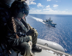 A Royal New Zealand Navy helicopter loadmaster looks out the SH-2G(I) Seasprite Helicopter door down to HMNZS Wellington sailing on the ocean on a nice day.