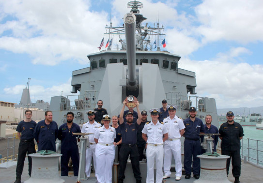 A group of Royal New Zealand Navy sailors stand on the front of the ship. One of the sailors is holding a trophy up above their head.