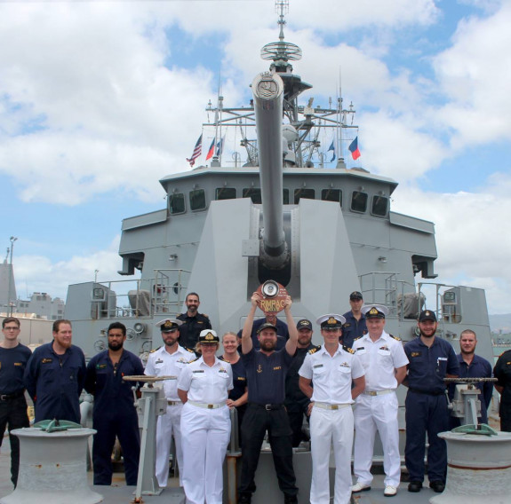 A group of Royal New Zealand Navy sailors stand on the front of the ship. One of the sailors is holding a trophy up above their head.