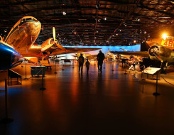 The inside of the Air Force Museum of New Zealand. The image shows aircraft in a darker display hanger and three people walking around the exhibit area.