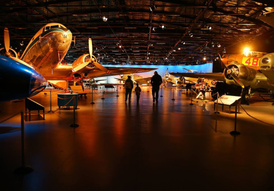 The inside of the Air Force Museum of New Zealand. The image shows aircraft in a darker display hanger and three people walking around the exhibit area.