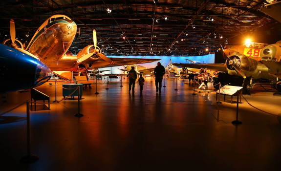 The inside of the Air Force Museum of New Zealand. The image shows aircraft in a darker display hanger and three people walking around the exhibit area.