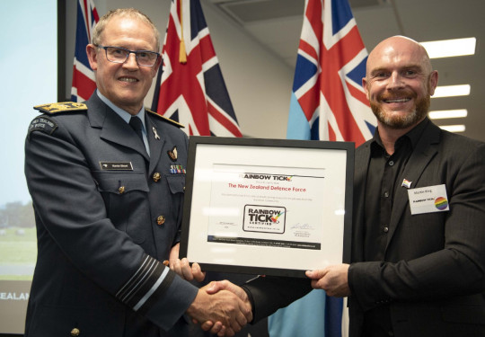 Chief of Defence Force Air Marshal Kevin Short and Martin King Director of the Rainbow Tick hold the Rainbow Tick certification and shake hands. The NZDF flags are in the background of the photo.