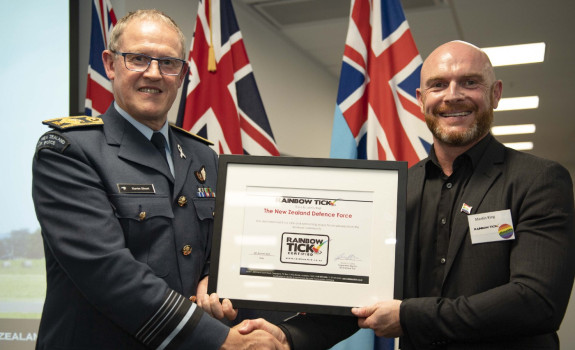 Chief of Defence Force Air Marshal Kevin Short and Martin King Director of the Rainbow Tick hold the Rainbow Tick certification and shake hands. The NZDF flags are in the background of the photo.