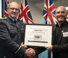 Chief of Defence Force Air Marshal Kevin Short and Martin King Director of the Rainbow Tick hold the Rainbow Tick certification and shake hands. The NZDF flags are in the background of the photo.