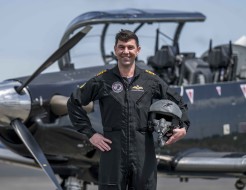 A military pilot smiles broadly whilst wearing a black flying suit. He stands in front of a small black aircraft with one hand on his hip and the other holding his helmet. 