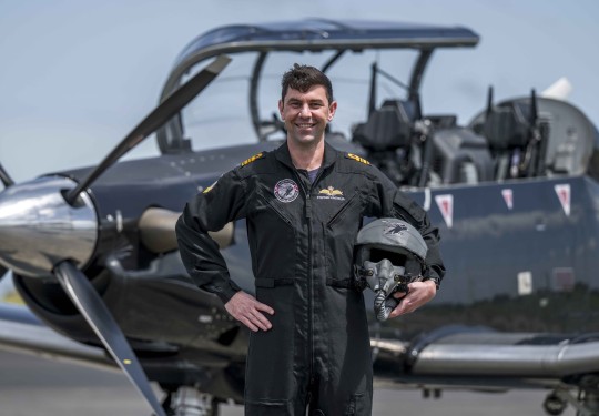 A military pilot smiles broadly whilst wearing a black flying suit. He stands in front of a small black aircraft with one hand on his hip and the other holding his helmet. 