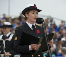 Able Musician Rebecca Nelson singing at a commemoration holding a folder with a microphone in front. in the background, out of focus you can see grounds sitting in stands and another Navy Band member.