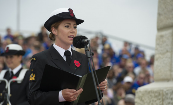 Able Musician Rebecca Nelson singing at a commemoration holding a folder with a microphone in front. in the background, out of focus you can see grounds sitting in stands and another Navy Band member.