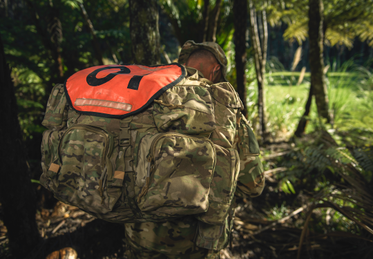 An SAS soldier walks through a tree lined path, his face hidden by his pack and hat.