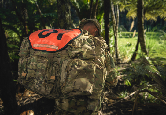 An SAS soldier walks through a tree lined path, his face hidden by his pack and hat.