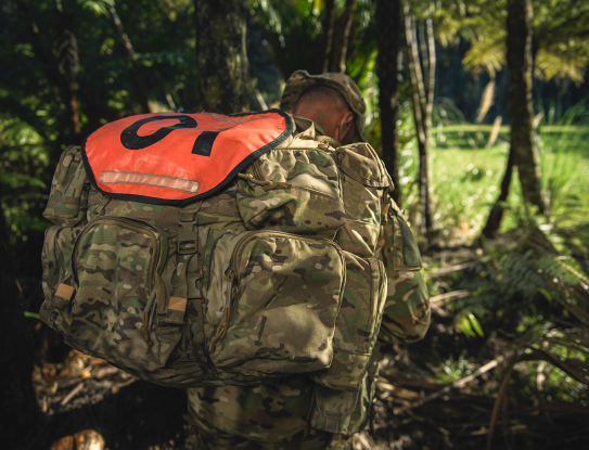 An SAS soldier walks through a tree lined path, his face hidden by his pack and hat.