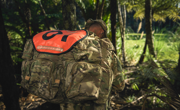 An SAS soldier walks through a tree lined path, his face hidden by his pack and hat.