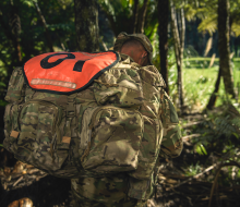 An SAS soldier walks through a tree lined path, his face hidden by his pack and hat.