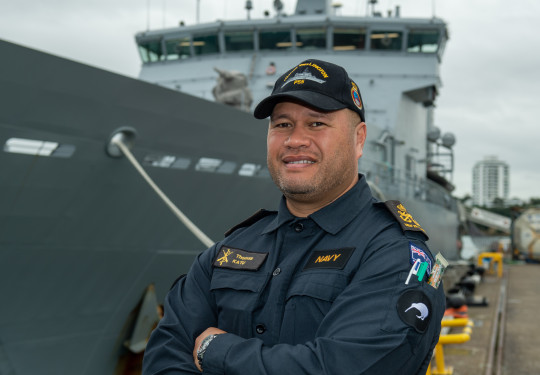 Sailor in working dress standing in front of an Offshore Patrol Vessel while ashore.