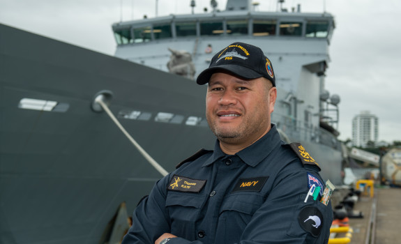 Sailor in working dress standing in front of an Offshore Patrol Vessel while ashore.