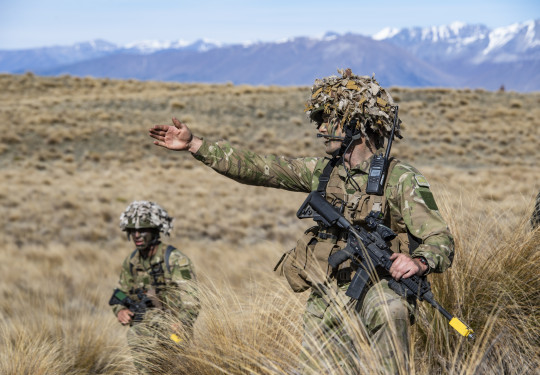 A soldier leads their section, pointing and directing to the right. Another soldier looks on, taking direction. The tussock and hill landscape of Tekapo stretches behind them.