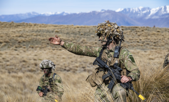 A soldier leads their section, pointing and directing to the right. Another soldier looks on, taking direction. The tussock and hill landscape of Tekapo stretches behind them.