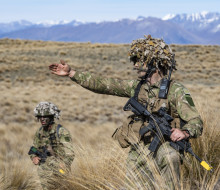 A soldier leads their section, pointing and directing to the right. Another soldier looks on, taking direction. The tussock and hill landscape of Tekapo stretches behind them.