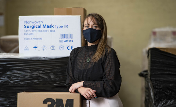 Tamara Hamiora is wearing a PPE mask, while leaning against a box of surgical masks and other PPE equipment.