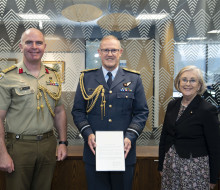 Three people stand in front of a glass wall with patterns on it. The New Zealand flag to the left. Chief of the New Zealand Defence Force Air Marshal Kevin Short (centre) was presented the commendation by Defence Adviser Colonel Neil Peake (left) and High