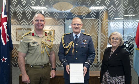 Three people stand in front of a glass wall with patterns on it. The New Zealand flag to the left. Chief of the New Zealand Defence Force Air Marshal Kevin Short (centre) was presented the commendation by Defence Adviser Colonel Neil Peake (left) and High