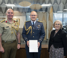 Three people stand in front of a glass wall with patterns on it. The New Zealand flag to the left. Chief of the New Zealand Defence Force Air Marshal Kevin Short (centre) was presented the commendation by Defence Adviser Colonel Neil Peake (left) and High