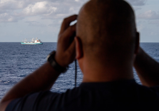 A sailor looks out of binoculars from the bridge of the ship to another boat.. 