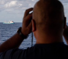 A sailor looks out of binoculars from the bridge of the ship to another boat..
