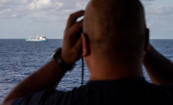 A sailor looks out of binoculars from the bridge of the ship to another boat..