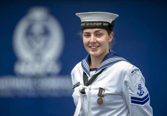 Leading Marine Technician (Propulsion) Jemma Hokai-Mataia looking at the camera in uniform with the Navy logo in background out of focus. 