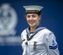 Leading Marine Technician (Propulsion) Jemma Hokai-Mataia looking at the camera in uniform with the Navy logo in background out of focus. 