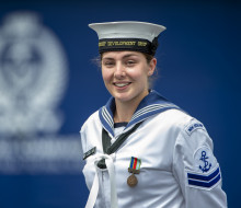 Leading Marine Technician (Propulsion) Jemma Hokai-Mataia looking at the camera in uniform with the Navy logo in background out of focus.