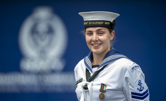 Leading Marine Technician (Propulsion) Jemma Hokai-Mataia looking at the camera in uniform with the Navy logo in background out of focus.