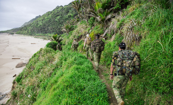 Soldiers trek in military gear with packs. A beach is to the left and hills stretch into the distance on the right.