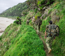Soldiers trek in military gear with packs. A beach is to the left and hills stretch into the distance on the right.