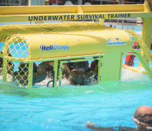 Soldiers in uniform and helmets are lowered into a pool by a crane, while sitting in a small metal helicopter simulator