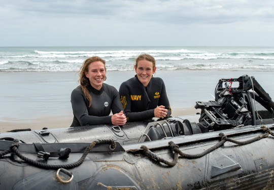 S1 Katrina Koch-Underhill (left) and S1 Sarah Gunderson (right) leaning on a Zodiac in front of the waves at Piha Beach.