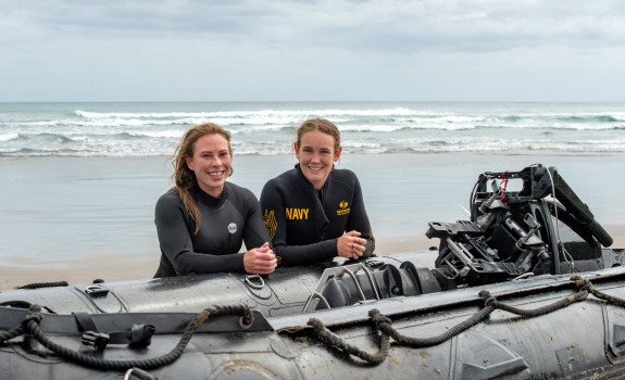 S1 Katrina Koch-Underhill (left) and S1 Sarah Gunderson (right) leaning on a Zodiac in front of the waves at Piha Beach.