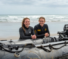 S1 Katrina Koch-Underhill (left) and S1 Sarah Gunderson (right) leaning on a Zodiac in front of the waves at Piha Beach.