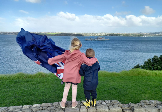 Family members wave farewell to Royal New Zealand Navy's HMNZS Aotearoa in Auckland. The two children are holding a flag and one of them has their arm around the other. 