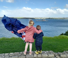 Family members wave farewell to Royal New Zealand Navy's HMNZS Aotearoa in Auckland. The two children are holding a flag and one of them has their arm around the other.