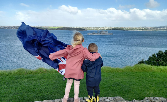 Family members wave farewell to Royal New Zealand Navy's HMNZS Aotearoa in Auckland. The two children are holding a flag and one of them has their arm around the other.