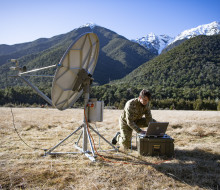 A Communications and Information Systems Mechanic student a radio systems in field conditions surrounded by tree-covered hills and snow-capped mountains.