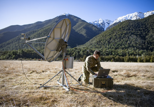 A Communications and Information Systems Mechanic student a radio systems in field conditions surrounded by tree-covered hills and snow-capped mountains.