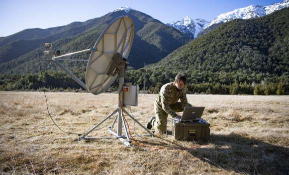 A Communications and Information Systems Mechanic student a radio systems in field conditions surrounded by tree-covered hills and snow-capped mountains.
