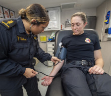 A woman wearing a dark blue Navy uniform with gold accents checks blood pressure of patient in a medical room.