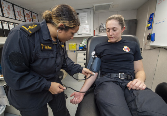 A woman wearing a dark blue Navy uniform with gold accents checks blood pressure of patient in a medical room.