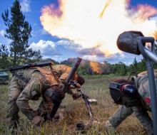 Three soldiers wearing hearing protection lower their heads as they fire a mortar, this creates a large flame . There are trees in the background and blue skies with scattered cloud.